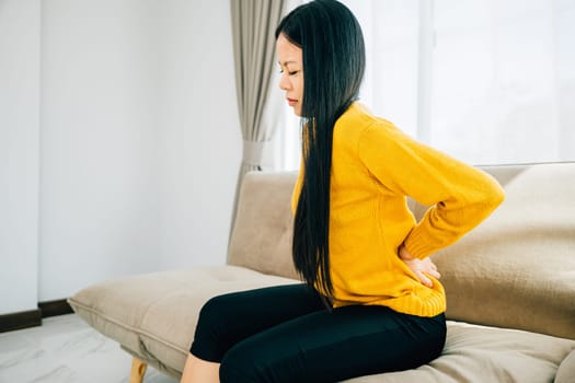 Depicting unbearable back pain, Asian woman seated on a sofa holds her lower back. Illustrating chronic backache discomfort and the necessity of medical care for back problems. health care concept