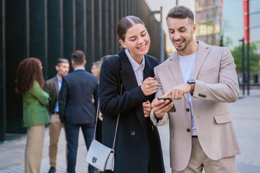 Two young business colleagues smiling while looking at a smartphone. They are standing outdoors in a modern urban setting, with other professionals in the background.