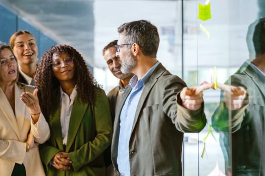 A diverse group of business professionals brainstorming in a modern office. A man points to sticky notes on a glass wall as the team collaborates with focus and engagement.