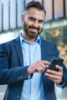 Smiling middle-aged businessman in a blue shirt holding and using a smartphone outdoors in a modern urban setting.