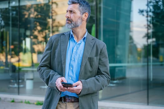 Middle-aged businessman in smart casual attire holding a smartphone, looking away thoughtfully in a modern urban environment.