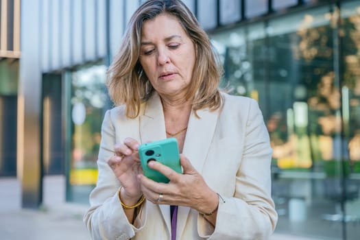 Smiling middle-aged businesswoman using a smartphone outdoors in a modern urban setting.