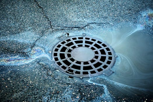 Gasoline creates colorful swirls on the water around a storm drain on wet asphalt.