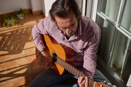 A man in casual clothing plays an acoustic guitar while seated on a patio. The natural sunlight adds warmth, underlining themes of relaxation, hobbies, and personal expression in an intimate outdoor setting.