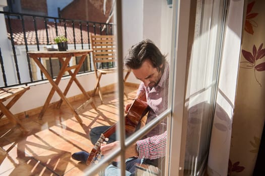 A man enjoys playing acoustic guitar while sitting on a sunny balcony. The terrace features wooden furniture, natural light, and a serene environment, making it a perfect scene for relaxation and music.