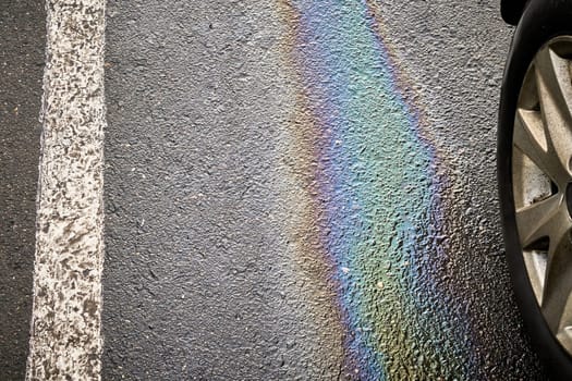 Colorful gasoline or oil spill creating a rainbow sheen on asphalt parking lot next to car tire and white parking line