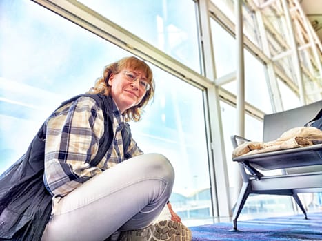 A middle-aged woman is seated in the airport terminal, taking a break and enjoying her surroundings as she awaits her upcoming flight.