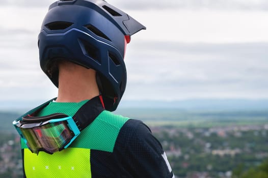 The back of a man in a protective helmet while he stands and looks at the city from the mountain. Professional racer, fully equipped with protective equipment, rests