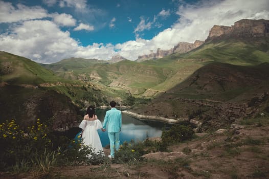 Romantic wedding couple in love standing on a rocky shore above a lake. Scenic view of majestic mountains and cloudy sky in summer.