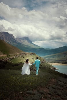 A wedding couple walks on green grass against the backdrop of mountains and high peaks. bride and groom stand with their backs turned and pose for wedding photos in the wilderness of the mountains.