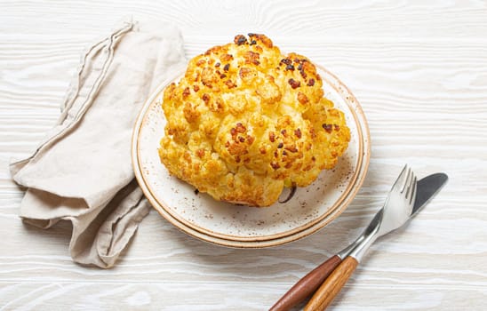 Whole roasted head of cauliflower with olive oil on plate on white wooden rustic table top view with cutlery. Vegetarian dish..