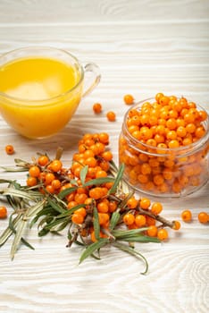 Sea buckthorn healthy hot tea in transparent glass cup, ripe berries in glass jar and branches with leaves on white wooden rustic background, great for skin, heart, vessels and immune system..