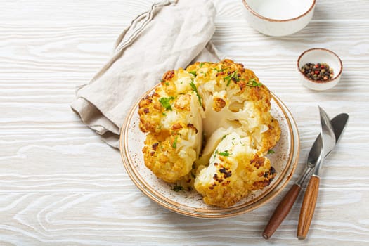 Whole roasted head of cauliflower with olive oil on plate on white wooden rustic table top view with cutlery. Vegetarian dish, space for text..