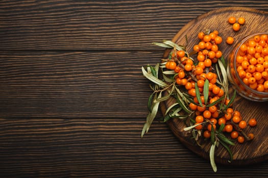 Sea buckthorn ripe berries in glass jar and branches with leaves top view on dark wooden rustic background, great for skin, heart, vessels and immune system. Copy space..