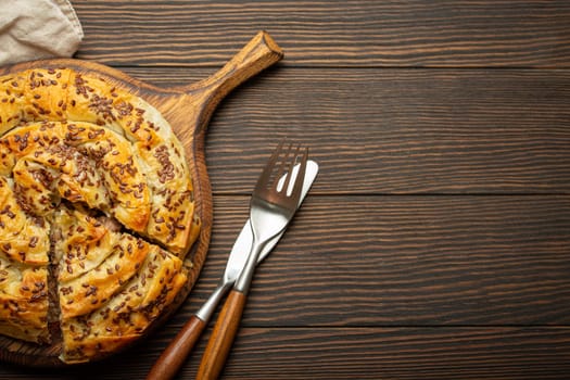 Burek made of filo dough with filling on cutting board, dark brown wooden rustic background top view. Traditional savoury spiral pie of Balkans, Middle East and Central Asia. Space for text.