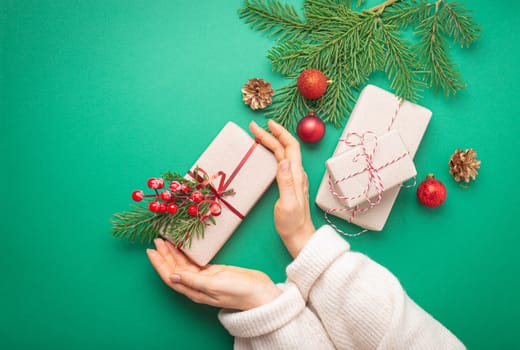 Christmas or New Year celebration green paper festive background with female hands holding wrapped present box, decoration fir tree, cones, berries, sparkly red balls. Space for text..