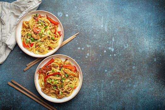 Two bowls with Chow Mein or Lo Mein, traditional Chinese stir fry noodles with meat and vegetables, served with chopsticks top view on rustic blue concrete background, space for text.