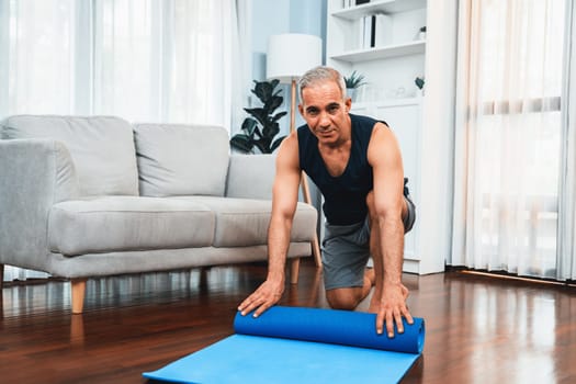 Active and sporty senior man preparing, rolling fitness exercising mat on living room floor at home. Home exercise as concept of healthy fit body lifestyle after retirement. Clout