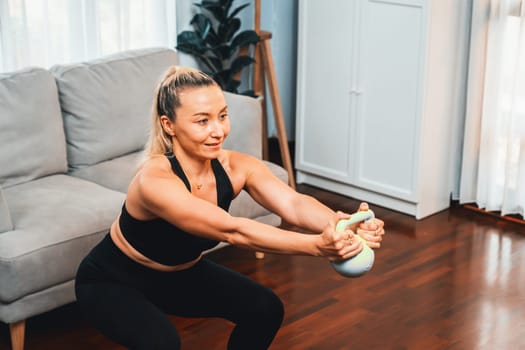 Athletic and sporty senior woman engaging in leg day training session with squat and bodyweight kettle ball at home exercise as concept of healthy fit body lifestyle after retirement. Clout