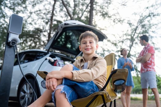 Little boy portrait sitting on camping chair with his family in background. Road trip travel with alternative energy charging station for eco-friendly car concept. Perpetual