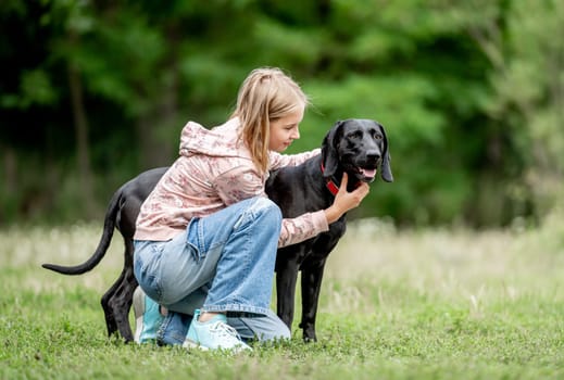 Preteen girl hugging golden retriever dog sitting at nature together. Cute child kid petting purebred pet doggy labrador in park at summer