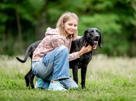 Preteen girl hugging golden retriever dog sitting at nature together. Cute child kid petting purebred pet doggy labrador in park at summer