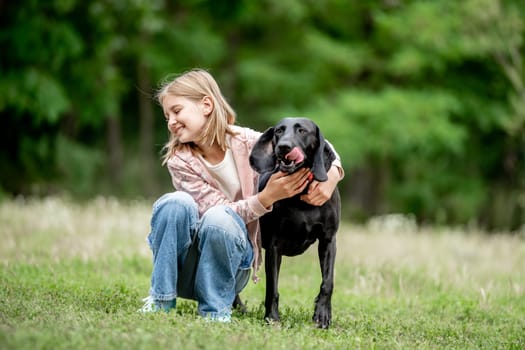 Preteen girl hugging golden retriever dog sitting at nature together. Cute child kid petting purebred pet doggy labrador in park at summer