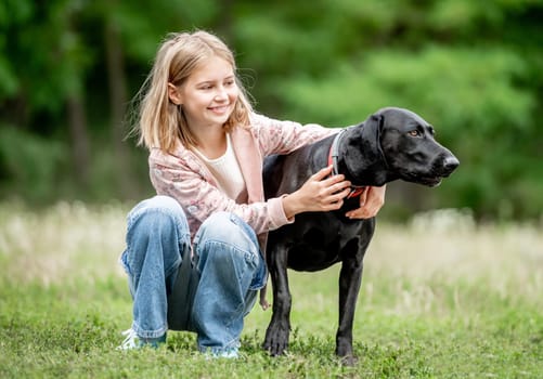 Preteen girl hugging golden retriever dog sitting at nature together. Cute child kid petting purebred pet doggy labrador in park at summer