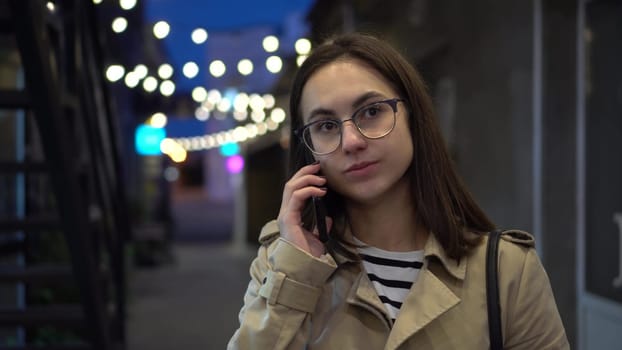 A young woman talks on the phone late at night on a narrow street. A girl with glasses communicates on a mobile phone. 4k