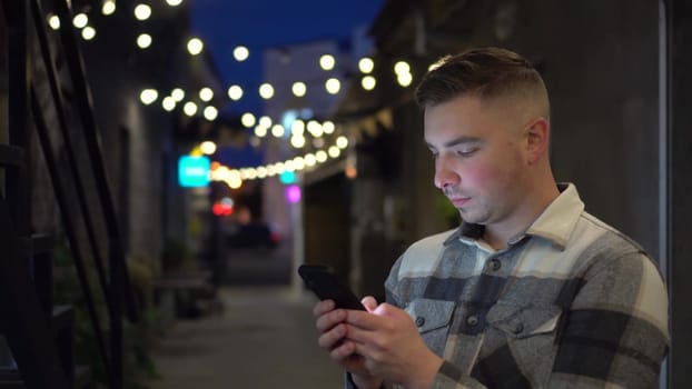 A young man chats on his smartphone late at night on a narrow street. A man in a warm shirt with a phone in his hands. 4k