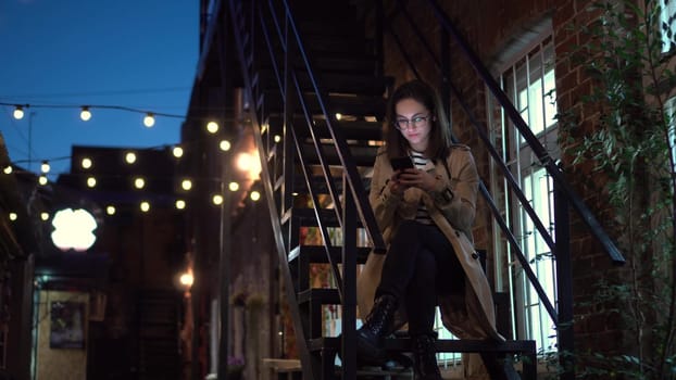 A young woman sits on a fire escape and texts on a smartphone. A girl with glasses on a narrow street late at night with a phone in her hands. 4k