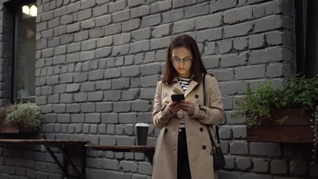 A young woman is chatting on a smartphone while standing against a wall on a narrow street. A girl in glasses and a polo with a phone in her hands. 4k