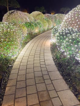 A winding stone path is surrounded by lush greenery and vibrant lights that create a magical atmosphere at night. The carefully arranged bushes are adorned with twinkling lights.