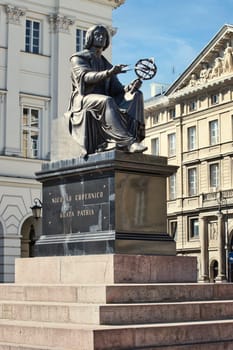 A detailed photograph of the Nicolaus Copernicus Monument in Warsaw, Poland. The statue portrays the renowned astronomer holding an armillary sphere, set against the elegant architecture of the surrounding buildings under a bright blue sky. Ideal for historical, educational, and travel themes.