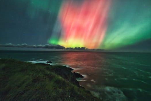 The ethereal Aurora Borealis lights up the night sky over the St. Abbs coastline in the UK. Shades of green and pink illuminate the scene, blending seamlessly with the dark, starry sky and reflecting on the ocean below. A captivating and serene depiction of the natural beauty of the northern lights, ideal for travel, nature, and celestial themes.