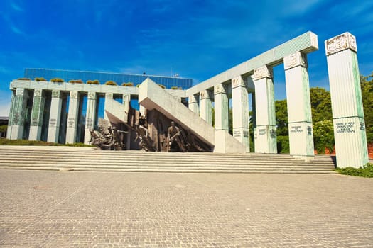 A powerful monument in Warsaw, Poland, commemorating the Warsaw Uprising of 1944. The dramatic sculpture features soldiers in action, surrounded by striking architectural elements and set against a bright blue sky. A poignant representation of courage, resilience, and history, ideal for historical and cultural themes.