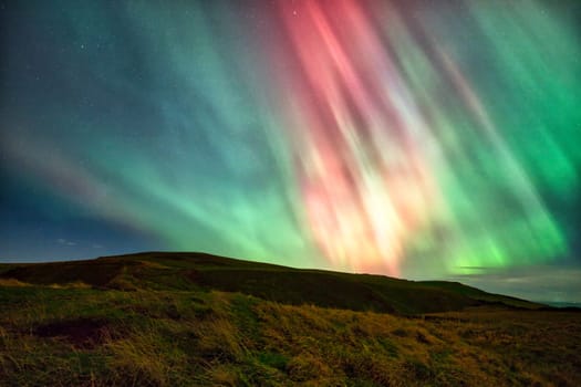 A breathtaking display of the Aurora Borealis, captured over the North Sea near St. Abbs, UK. The vibrant green and pink lights of the northern lights stretch across the night sky, reflecting beautifully on the water. The foreground features grassy cliffs, adding depth and contrast to the serene yet dramatic scene. Perfect for travel, nature, and astronomy themes.