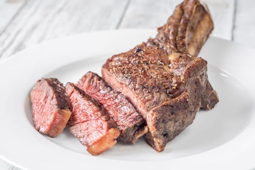 Sliced grilled medium-rare cowboy steak served on a white plate atop a white wooden table