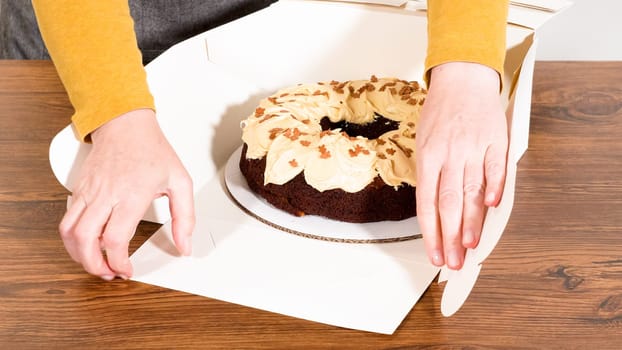Carefully placing the gingerbread bundt cake, adorned with salted caramel frosting and gingerbread sprinkles, into a white paper bundt cake box with a clear window for gifting.