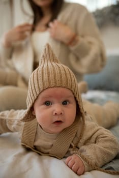 A baby wearing a hat. A small child is crawling on a bed. His mother is sitting behind him. The little boy is wearing a knitted beige hat. the baby is very active and cute, he crawls on the bed.