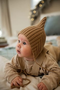 A baby wearing a hat. A small child is crawling on a bed. His mother is sitting behind him. The little boy is wearing a knitted beige hat. the baby is very active and cute, he crawls on the bed.