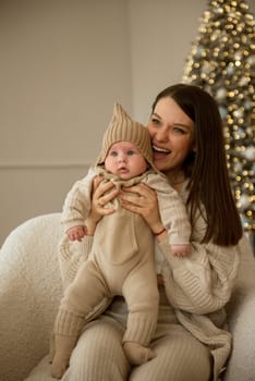 A woman holding a baby.The girl holds her child in her arms. The child is wearing a beige hand-knitted hat. mother and child are happy, they are smiling. They celebrate Christmas.