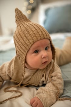 A baby wearing a hat. A small child is crawling on a bed. His mother is sitting behind him. The little boy is wearing a knitted beige hat. the baby is very active and cute, he crawls on the bed.