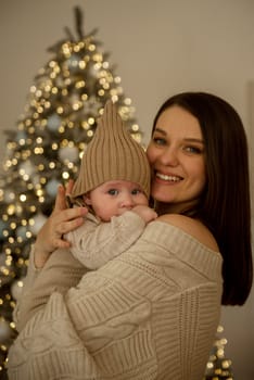 A woman holding a baby.The girl holds her child in her arms. The child is wearing a beige hand-knitted hat. mother and child are happy, they are smiling. They celebrate Christmas.