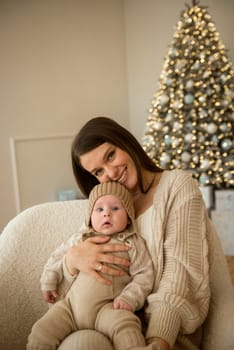 A woman holding a baby.The girl holds her child in her arms. The child is wearing a beige hand-knitted hat. mother and child are happy, they are smiling. They celebrate Christmas.