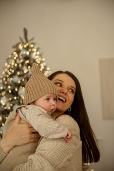 A woman holding a baby.The girl holds her child in her arms. The child is wearing a beige hand-knitted hat. mother and child are happy, they are smiling. They celebrate Christmas.