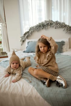The boy is small, he is lying on his stomach. His sister, a girl of about 7 years old, is sitting next to him. They are in the bedroom on the bed.the children are dressed in beige clothes