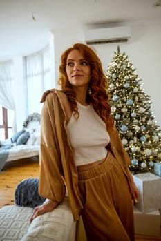 A woman sitting on a bed.A girl with red curly hair is sitting near the bed in the bedroom. Behind her in the room is a Christmas tree with toys and gifts. The girl is dressed in white knitted clothes