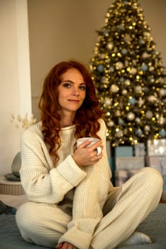 A woman sitting on a bed.A girl with red curly hair is sitting near the bed in the bedroom. Behind her in the room is a Christmas tree with toys and gifts. The girl is dressed in white knitted clothes