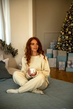 A woman sitting on a bed.A girl with red curly hair is sitting near the bed in the bedroom. Behind her in the room is a Christmas tree with toys and gifts. The girl is dressed in white knitted clothes